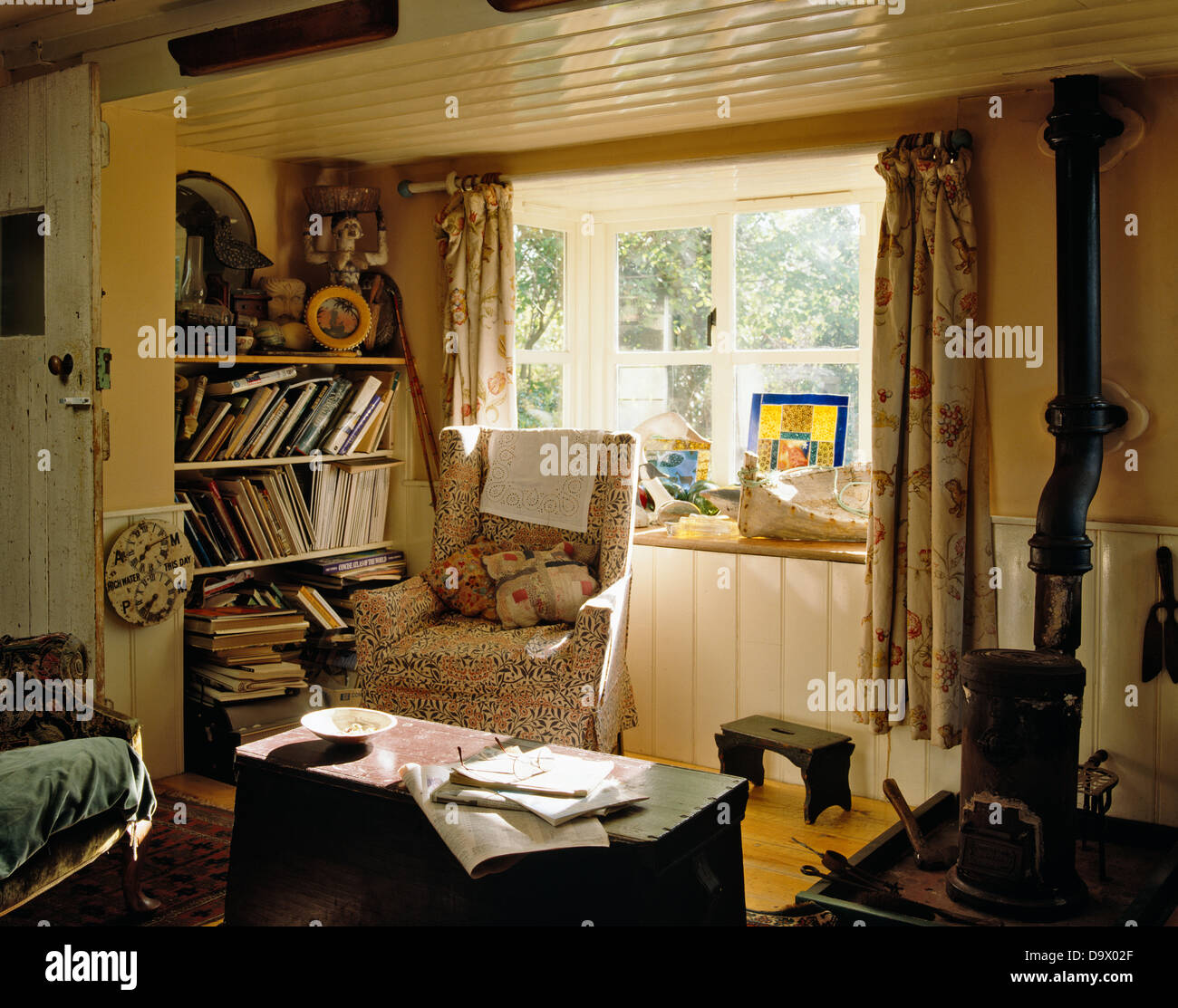 Poltrona davanti alla finestra in basso soffitto cottage soggiorno con scaffali disordinato e la piccola stufa a legna Foto Stock