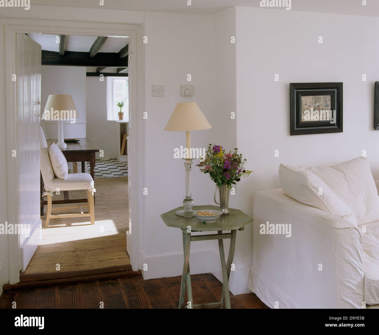 La crema lampada dipinta sul tavolino accanto al divano bianco in bianco cottage soggiorno con porta aperta alla cucina Foto Stock