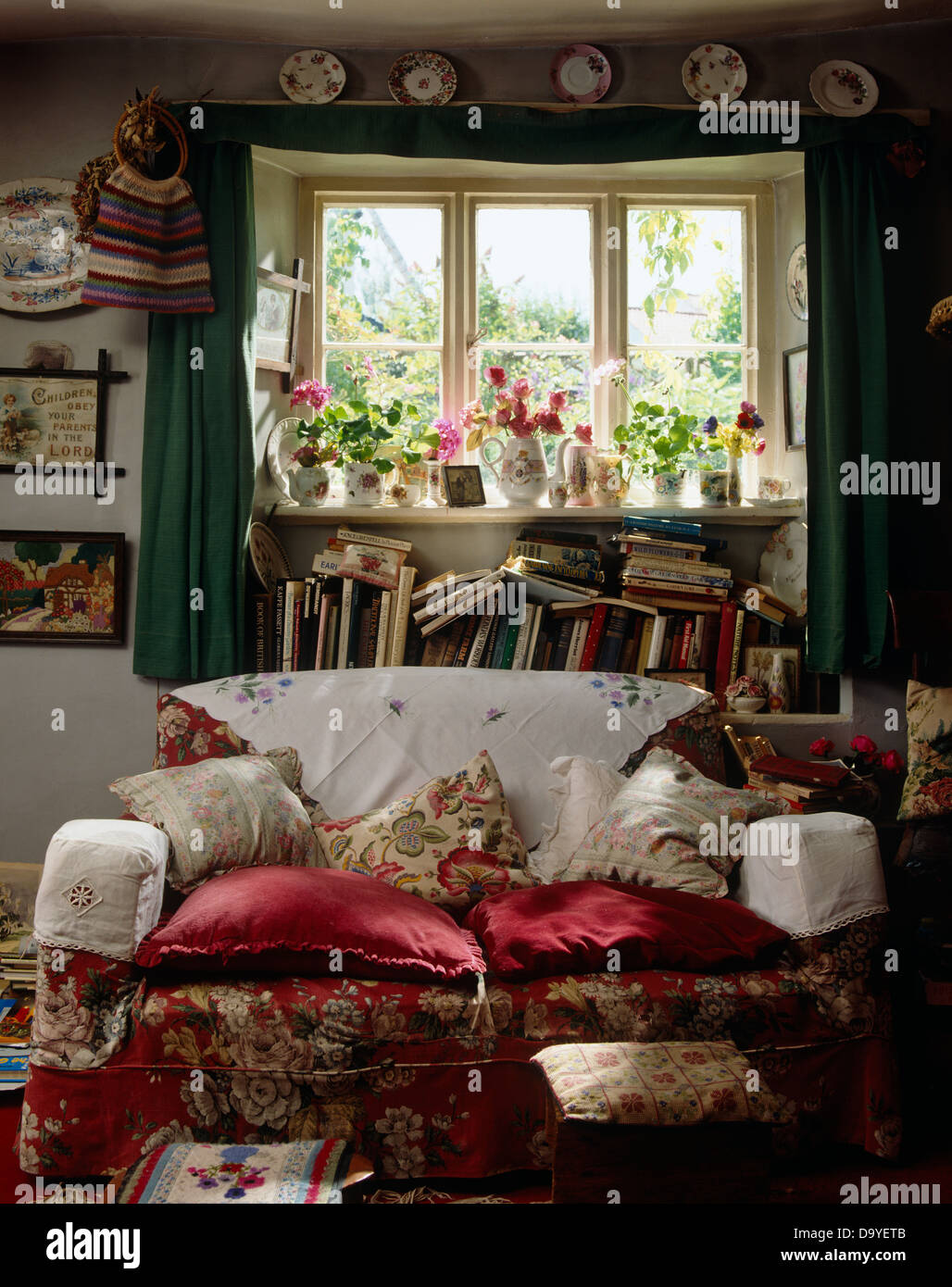 Antimacassars bianco e cuscini rossi sul divano floreale in cottage ingombra la stanza di seduta con la pila di libri disordinata finestra sottostante Foto Stock