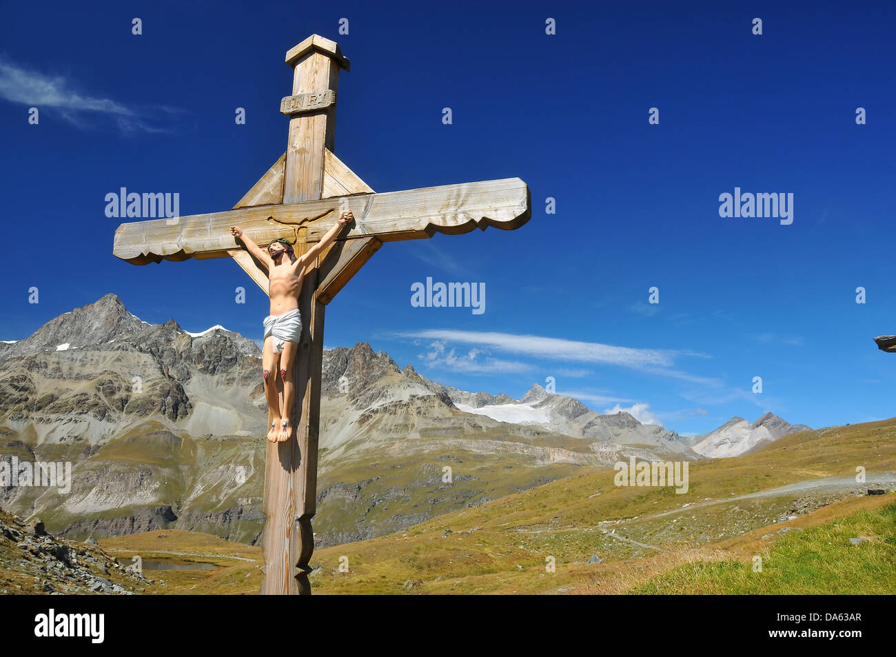 Croce con Gesù Cristo crocifisso, in montagna, al di sopra di un lago, con alte alpi nella distanza, in una limpida giornata di blu Foto Stock