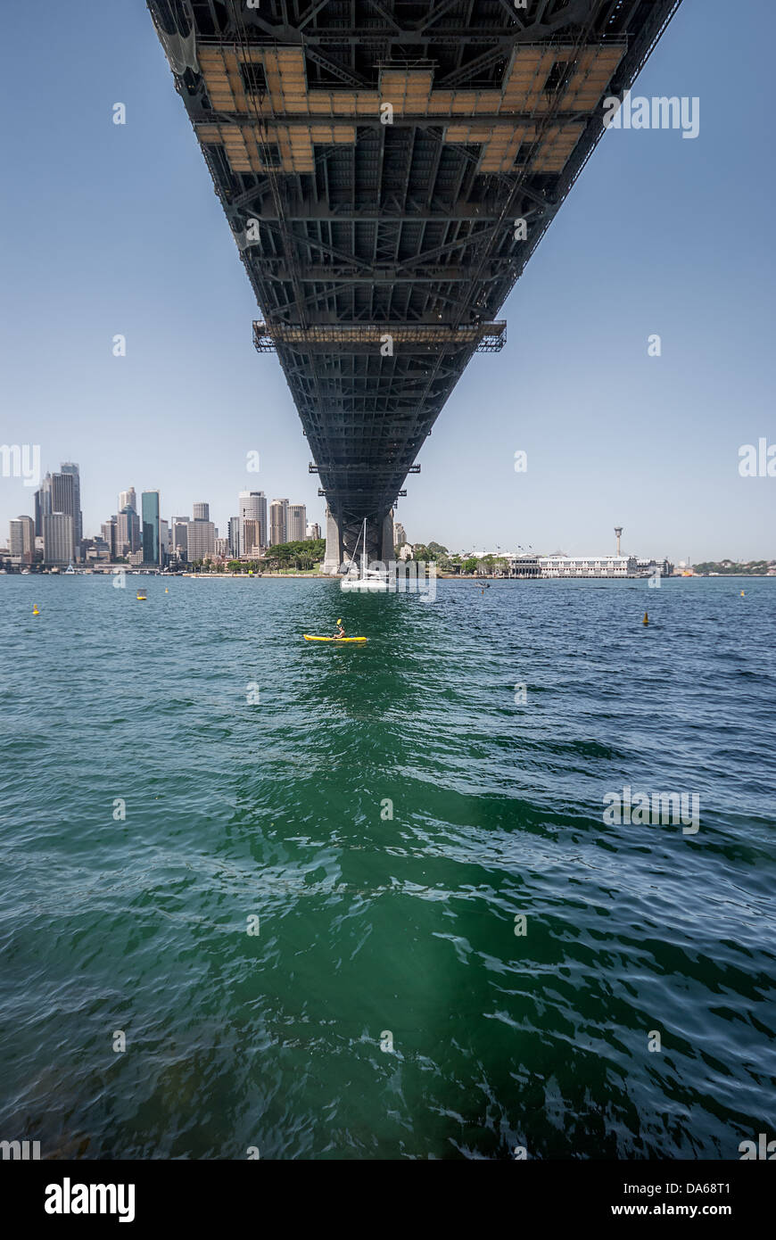Una vista unica di una barca a vela, nanizzata dalla parte inferiore dell'iconico Sydney Harbour Bridge, che si estende dal centro a North Sydney. Foto Stock