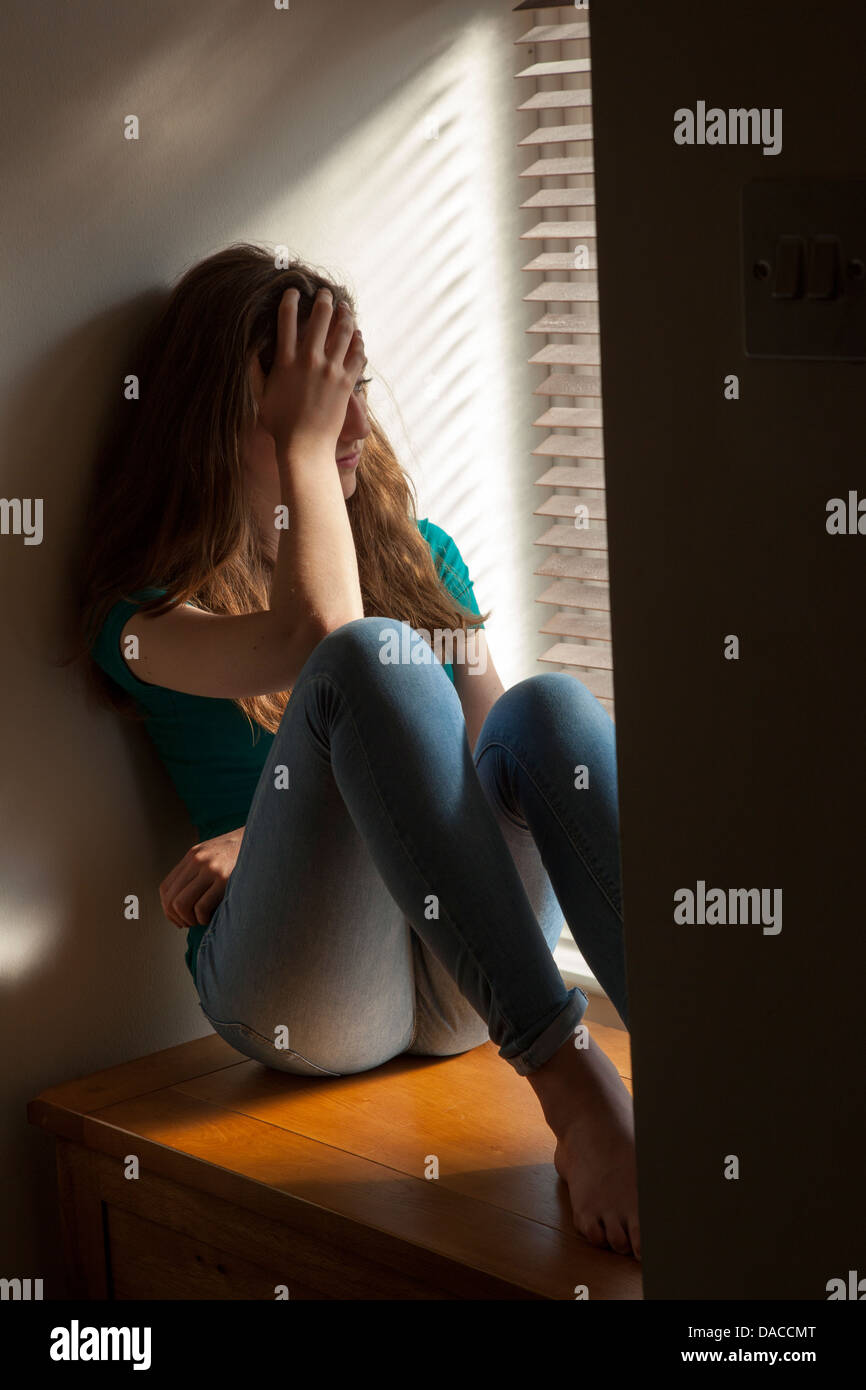Una ragazza adolescente, la mano sulla sua testa, seduti in una finestra con una luce di colata. Femmine giovane adulto Foto Stock