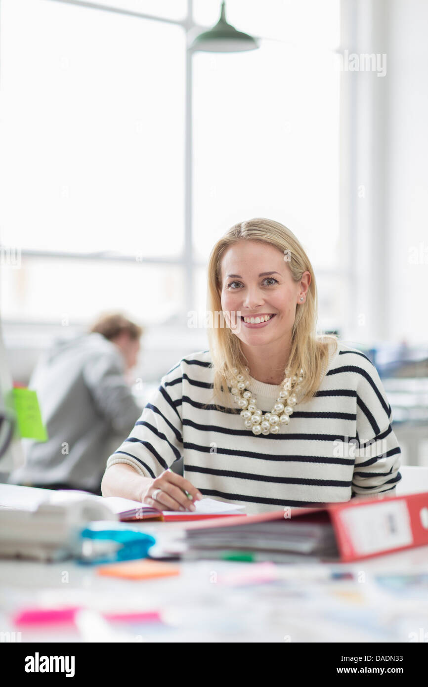 Giovane donna seduta alla scrivania e sorridente in ufficio creativo, ritratto Foto Stock