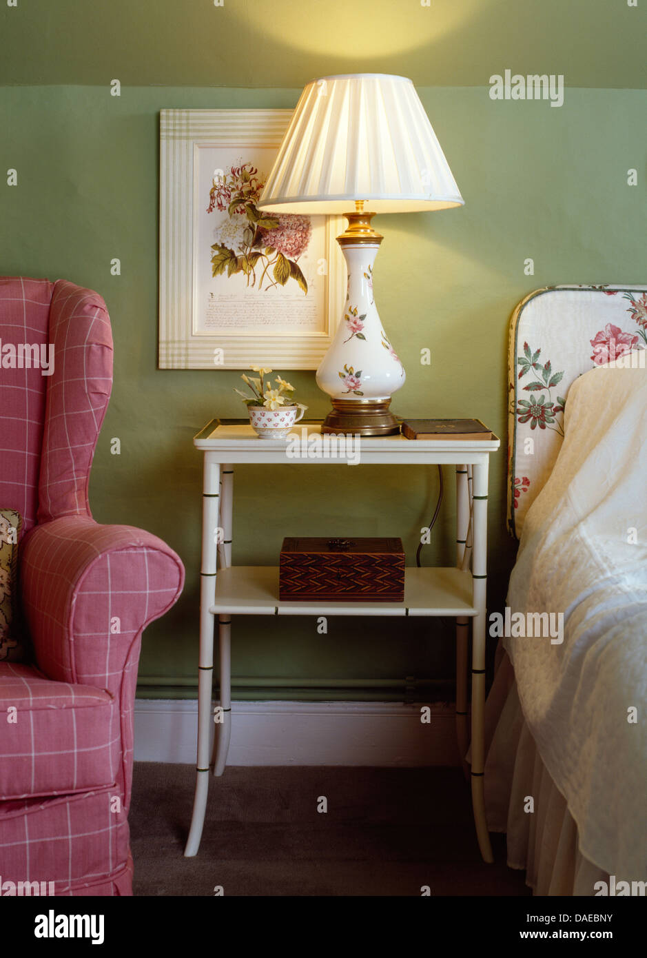 Illuminato lampada floreale dipinta sul comodino tra letto e verificato rosa poltrona in verde cottage camera da letto Foto Stock
