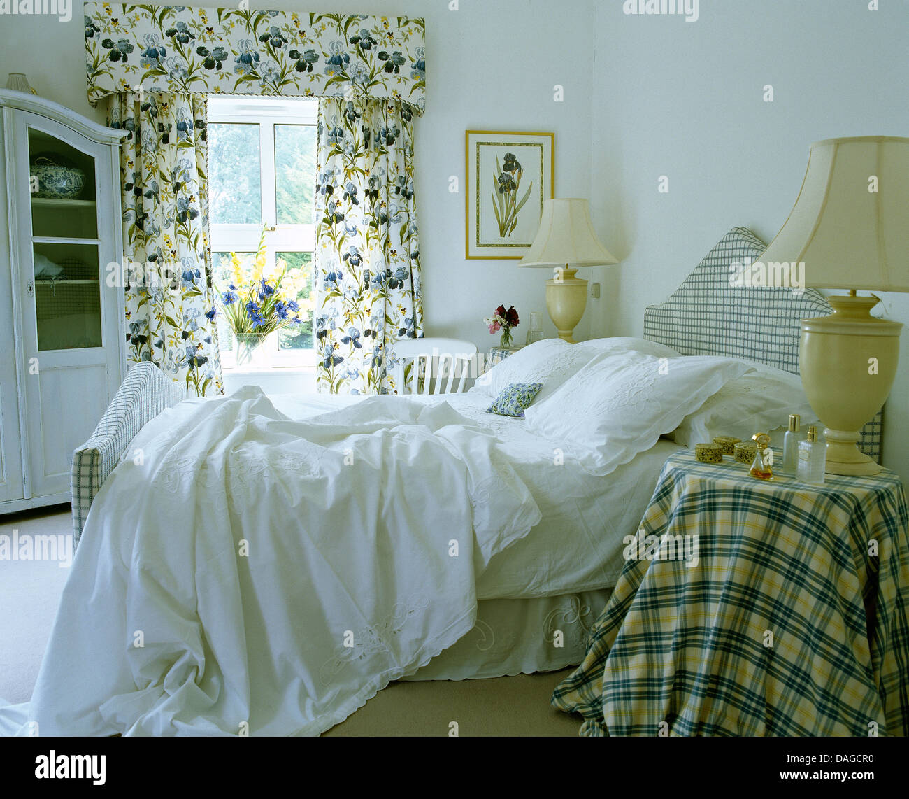 Cuscini bianchi e fogli spiegazzati sul letto nella camera da letto del paese controllato con panno sul comodino e drappeggi floreali sulla finestra Foto Stock