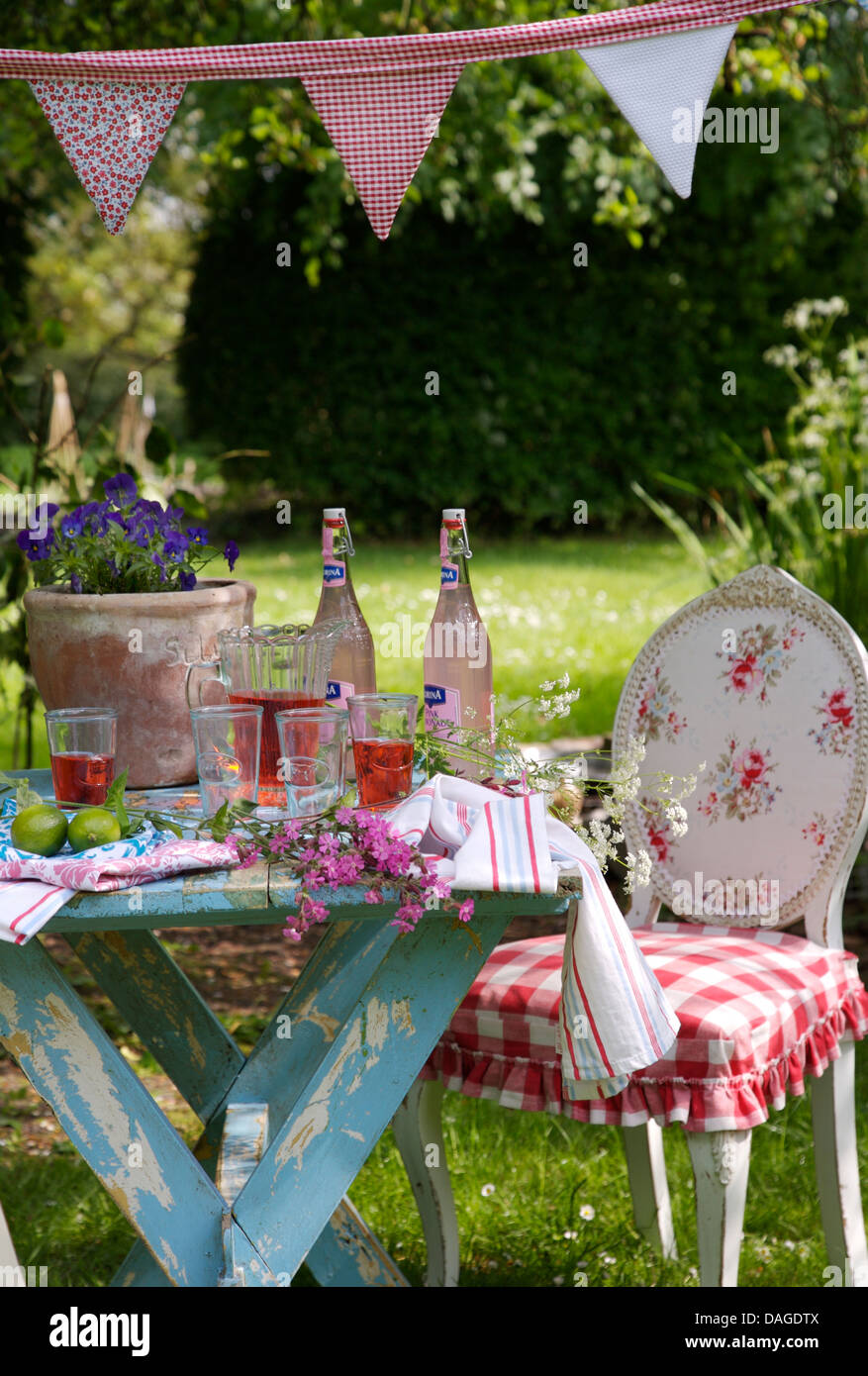 Giardino estivo con dipinto di blu tabella impostata per il pranzo con le bottiglie e i bicchieri di frutta rosa cordiale Foto Stock