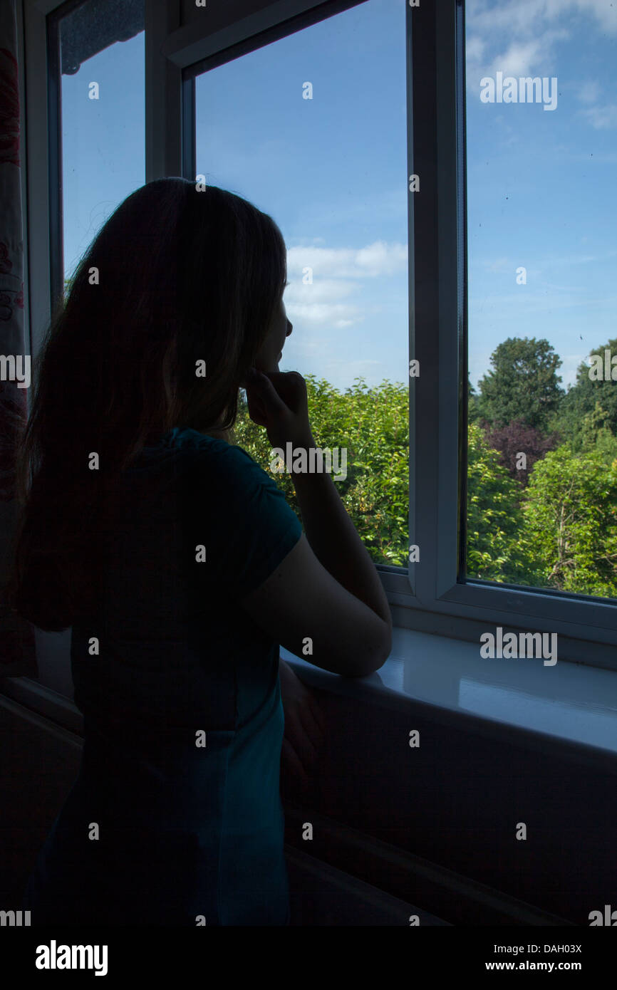 Ragazza adolescente in silhouette guardando fuori della sua camera da letto finestra. Foto Stock
