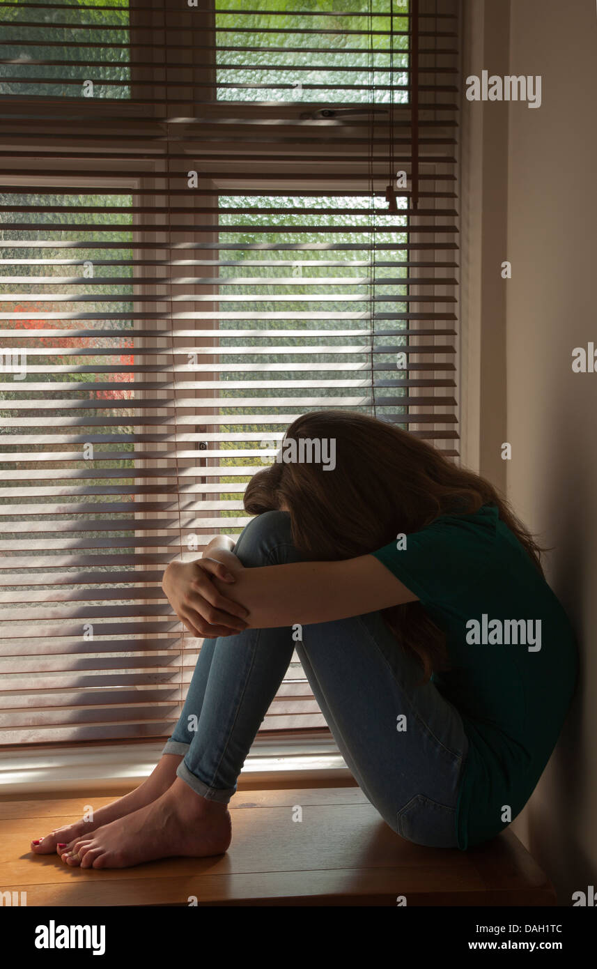 Una ragazza adolescente, la testa tra le ginocchia, seduti in una finestra con una luce di colata. Foto Stock