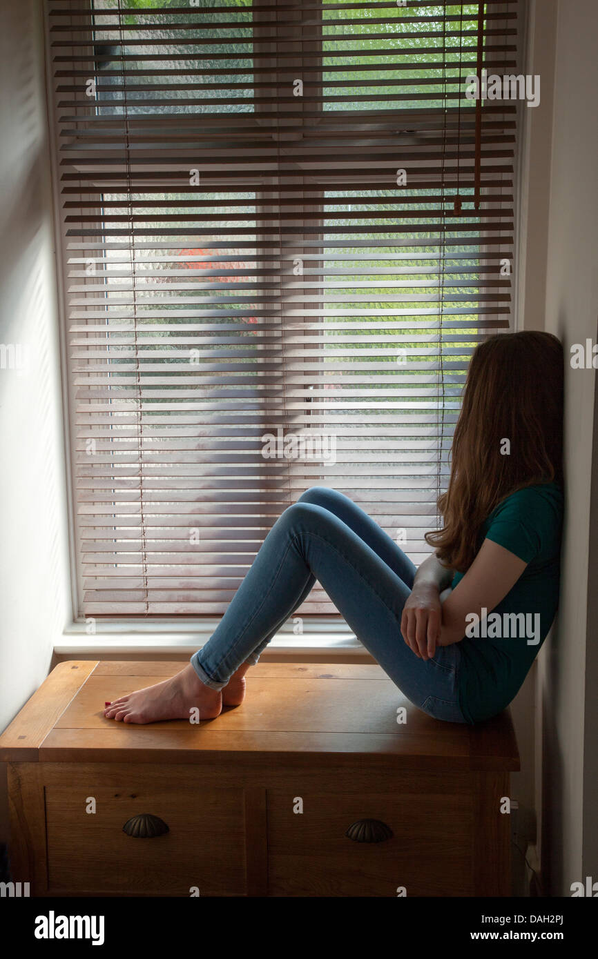 Una ragazza adolescente, seduti nelle tenebre da una finestra con la luce all'esterno. Foto Stock