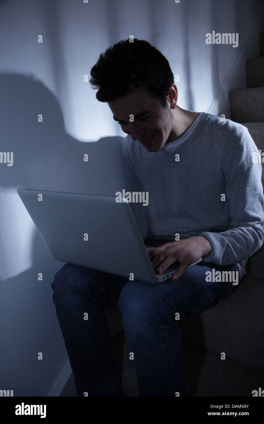 Maschio adolescente usando il suo computer portatile, seduta in scala scura.. Foto Stock