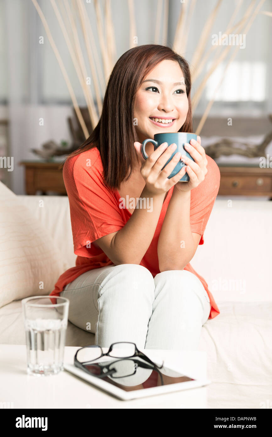 Bella donna cinese tenendo una tazza di caffè durante la seduta sul suo divano Foto Stock