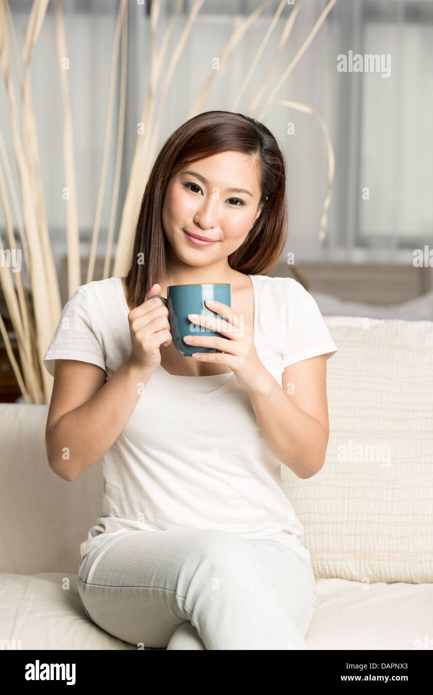 Bella donna cinese tenendo una tazza di caffè durante la seduta sul suo divano Foto Stock
