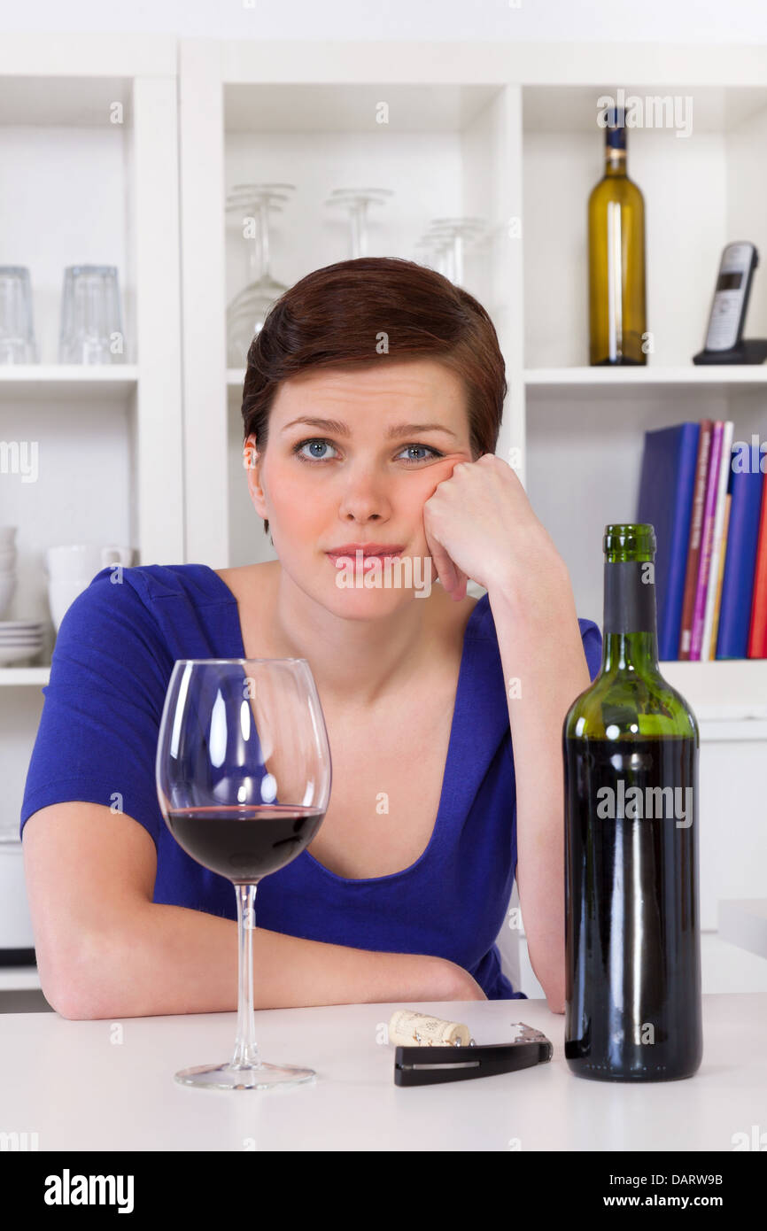 Giovane triste thinkful donna seduta con un bicchiere di vino rosso nella sua cucina Foto Stock
