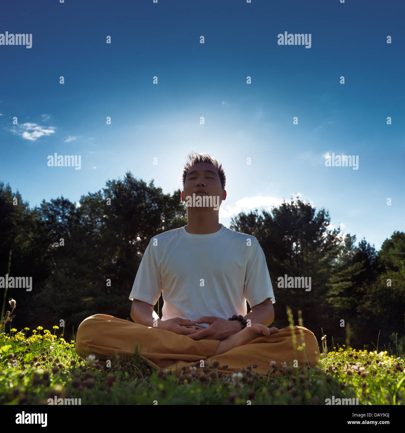 Uomo cinese meditando all'aperto durante l'alba nella natura, seduta con le gambe incrociate su erba sotto il cielo blu Foto Stock