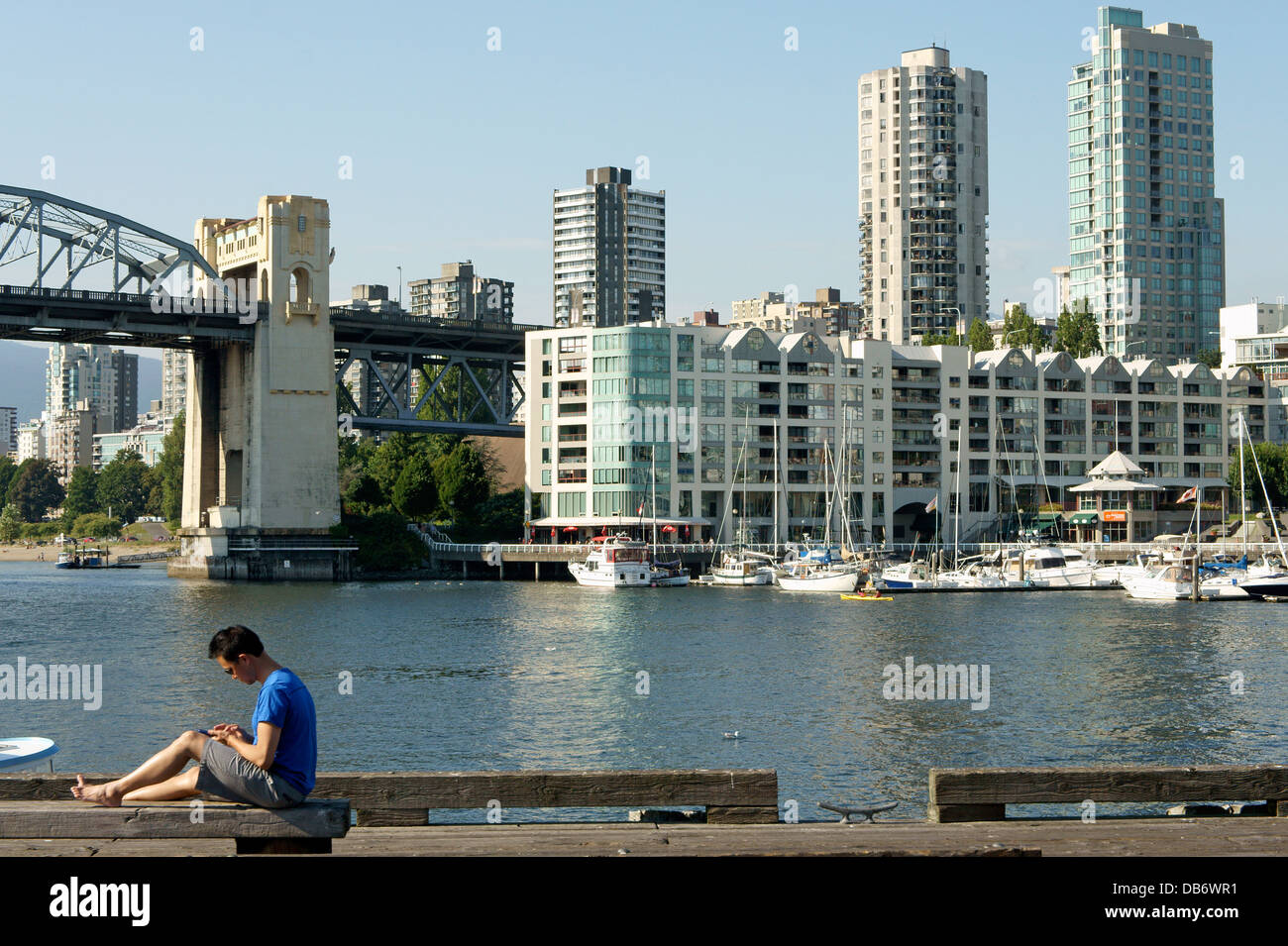 Giovane uomo asiatico controllando il suo smart phone mentre è seduto su di un molo, Granville Island, Vancouver, British Columbia, Canada Foto Stock