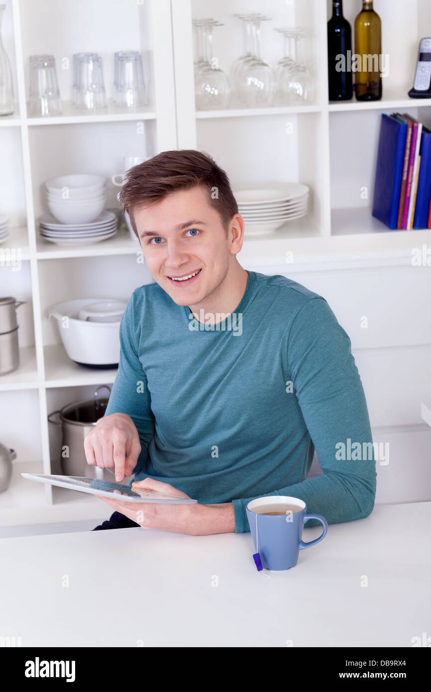 Giovane uomo seduto con un tablet PC e una tazza di tè in cucina Foto Stock