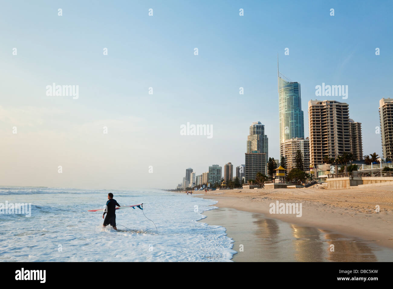 Surfer a piedi in surf con skyline della città in background. Surfers Paradise, Gold Coast, Queensland, Australia Foto Stock