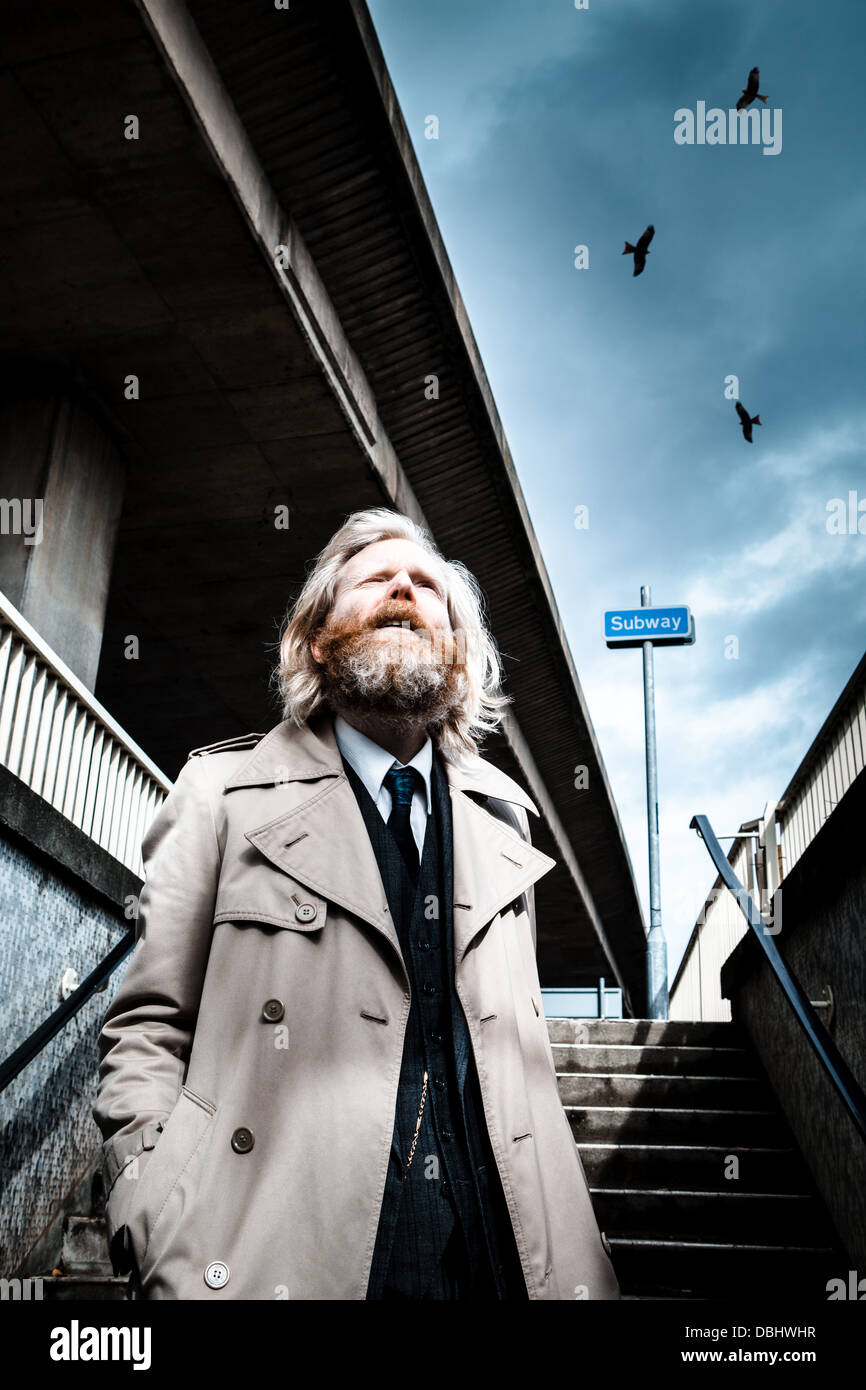 Uomo Barbuto in tuta e soprabito passeggiando per le fasi in una metropolitana, minacciosi uccelli overhead. Foto Stock