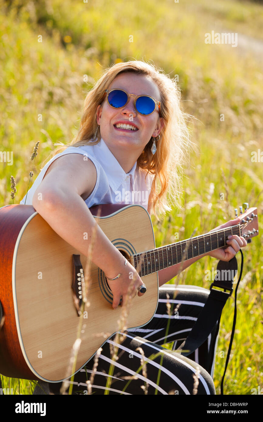 Ragazza adolescente indossando occhiali scuri a suonare la chitarra in un campo erboso. All'esterno. L'estate. Foto Stock