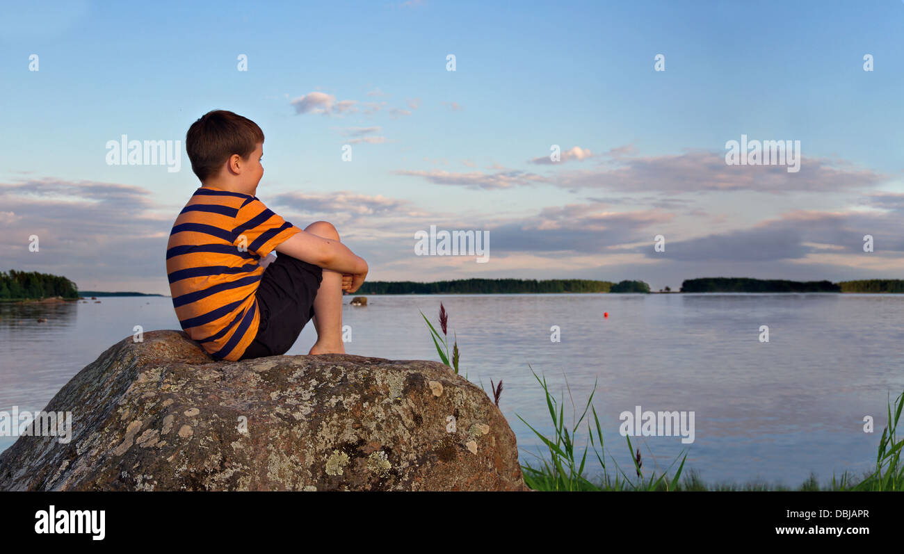 Panorama di un giovane ragazzo seduto su una grande roccia in una serata estiva alla luce del sole la visione di un paesaggio balneare Foto Stock