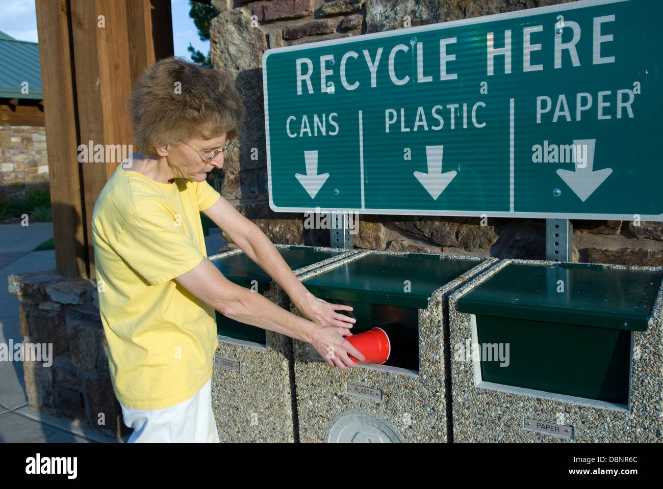 Donna caucasica che lancia una tazza di plastica nel bidone di riciclaggio verde in una giornata di sole negli Stati Uniti, promuovendo la responsabilità ambientale. Foto Stock