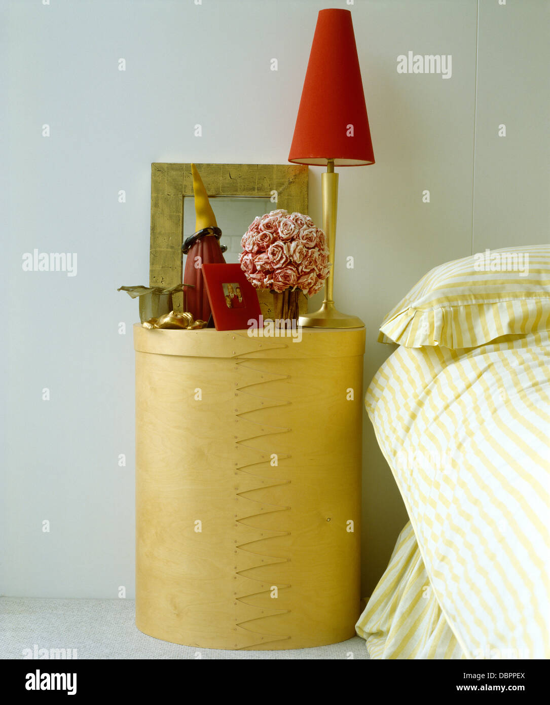 Lampada rossa e vaso di rose rosa su cilindrica del tamburo di legno compensato tavolo accanto a letto con il giallo listati piumone Foto Stock