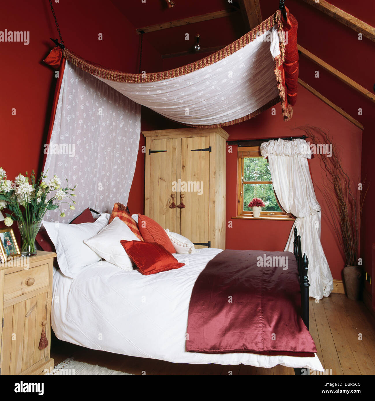 Tettoia con voile bianco rivestimento sopra il letto con il piumone bianco e raso rosso buttare in rosso mansarda camera da letto in cottage di campagna Foto Stock
