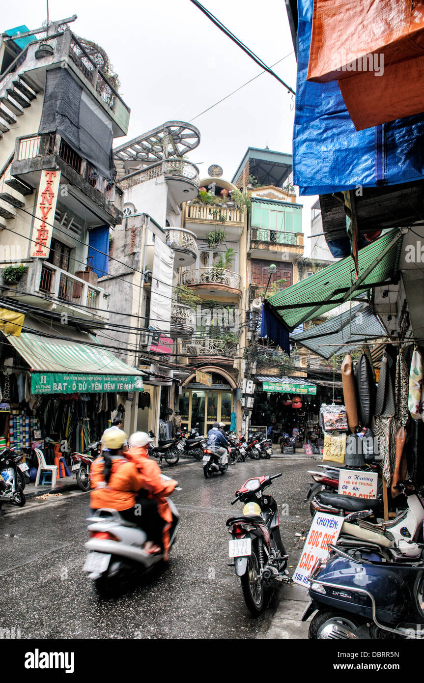 Market Street Tube Houses Motorbikes Wet Pavement Hanoi Vietnam | HANOI, Vietnam — Motociclette navigano su un marciapiede bagnato attraverso un distretto commerciale incorniciato da edifici stretti e multipiano e da una fitta rete di cavi aerei. Queste strutture distintive, conosciute come nhà ống o case tubo, dominano il quartiere storico della città. La loro estrema strettezza è nata in risposta alle tasse immobiliari storiche calcolate in base al fronte stradale, spingendo i residenti a costruire in profondità e in altezza piuttosto che in larghezza, producendo case-negozio che tipicamente misurano solo da 6 a 13 piedi (2 a 4 m) di larghezza ma si estendono per decine di metri verso il retro Foto Stock