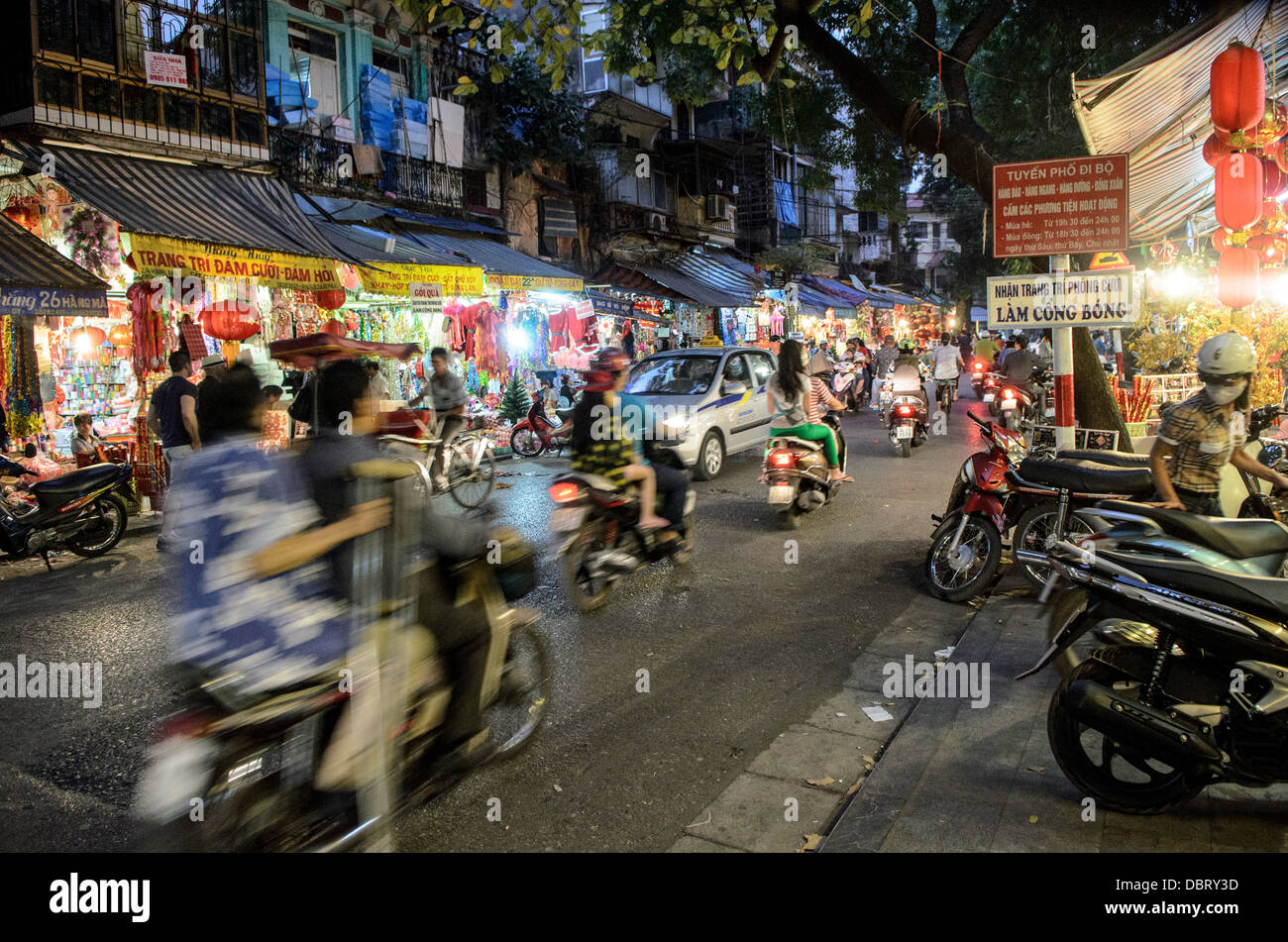 Quartiere Antico Sfocato Scooter Lanterne Rosse Hanoi Vietnam | HANOI, Vietnam — Le moto sfrecciano davanti alle vetrine dei negozi piene di lanterne su Hàng Mã, una delle storiche “36 strade” del Quartiere Antico, chiamata così per i prodotti di carta votiva che la sua gilda un tempo vendeva. Le strade qui erano tradizionalmente prefissate con “Hàng” (“merce”) per il commercio specifico praticato lungo di esse, un sistema di denominazione che risale alla specializzazione delle corporazioni artigiane sotto la dinastia Lê. Foto Stock