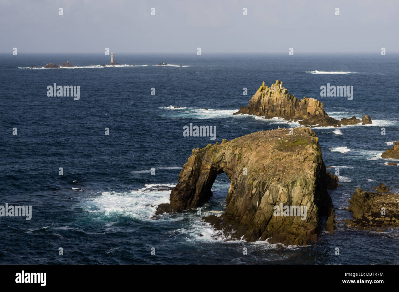 Un paesaggio marino di formazioni rocciose e un faro della distanza appena fuori il paese di costa di estremità in Cornovaglia. Foto Stock