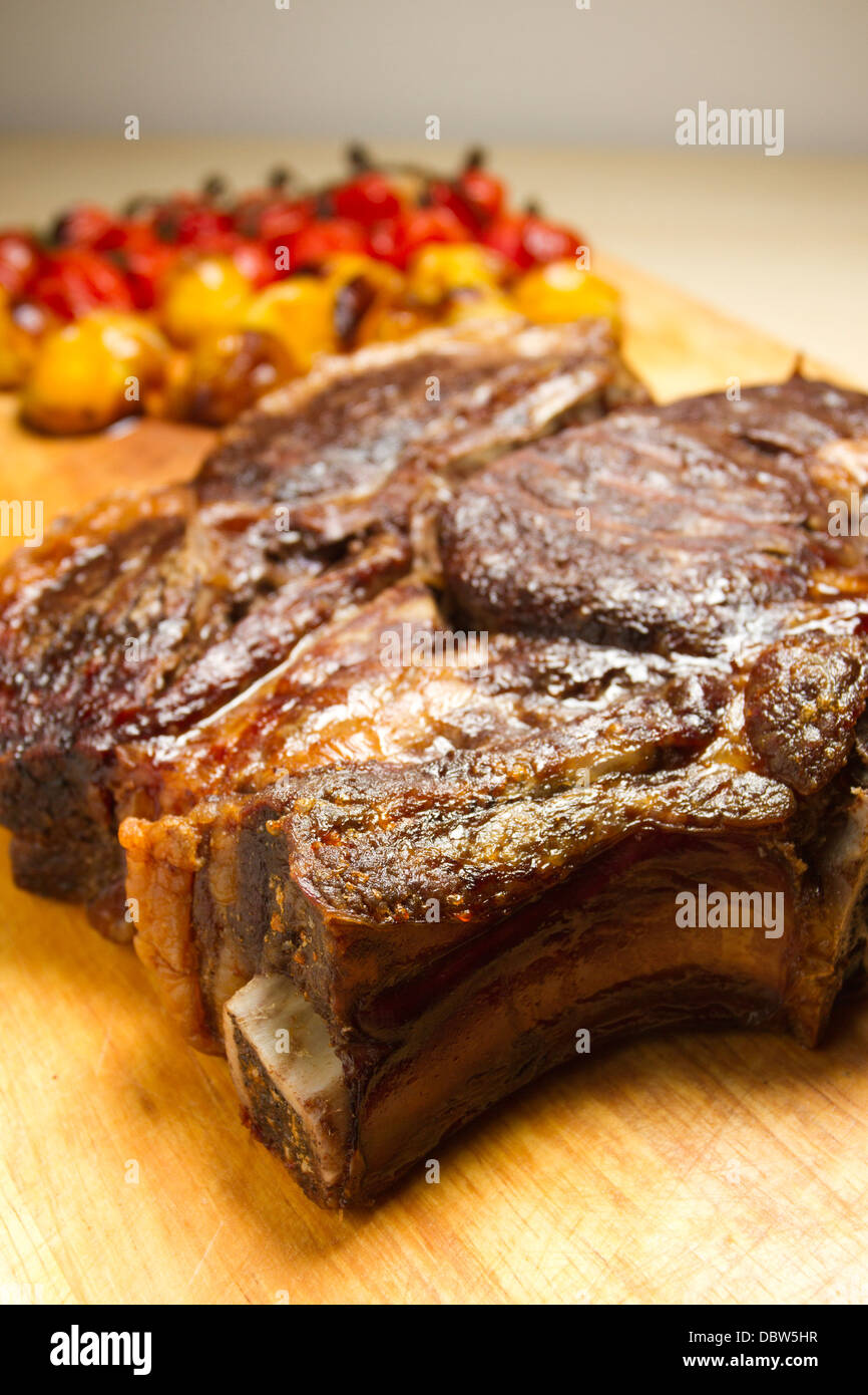 Costola arrosto di carne su un tagliere di legno, con arrosti di carne rossa e pomodori giallo Foto Stock