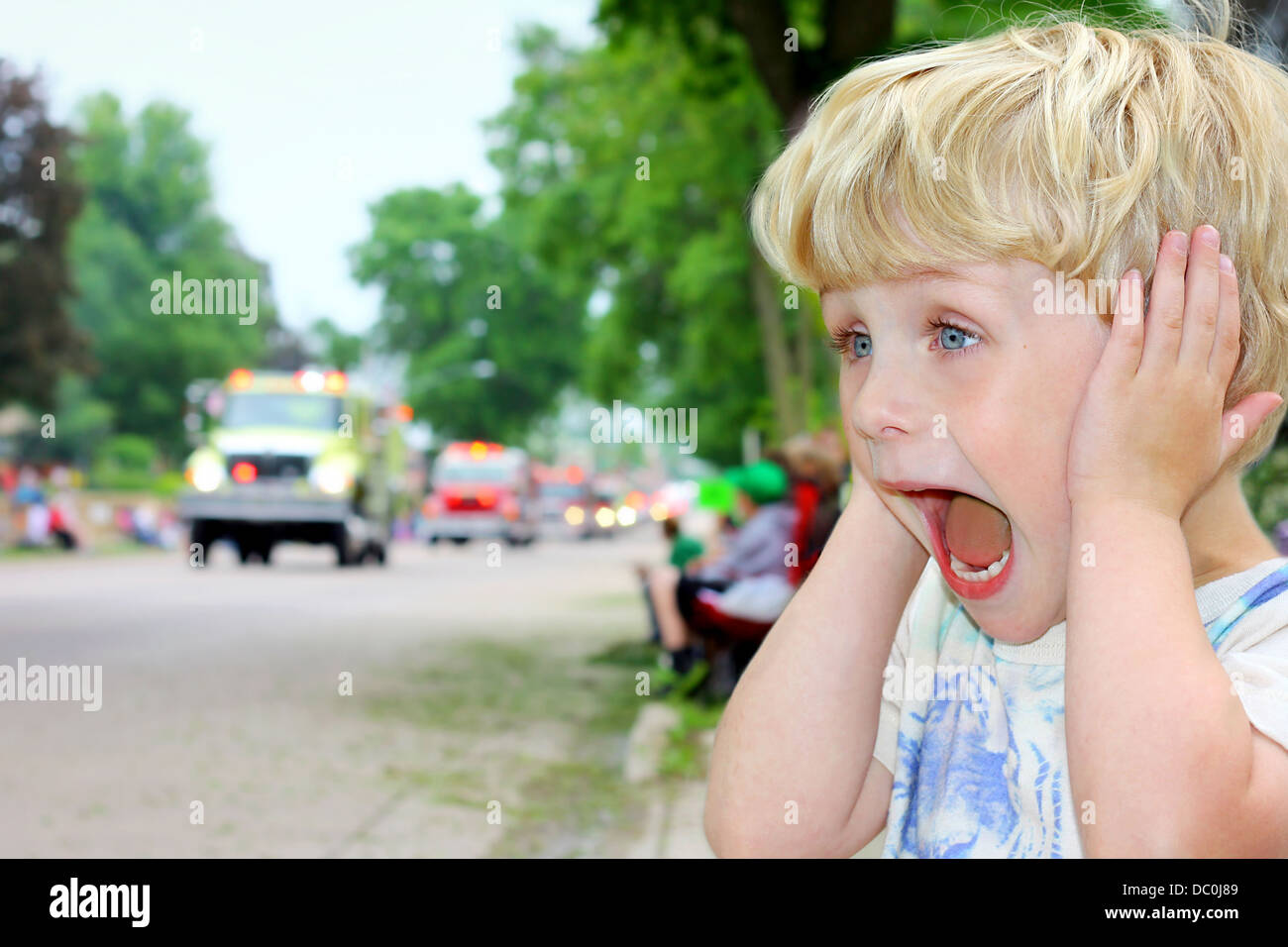 Un giovane ragazzo biondo copre le orecchie e guarda eccitato come ambulanze e camion fuoco guidare da in una parata. Foto Stock