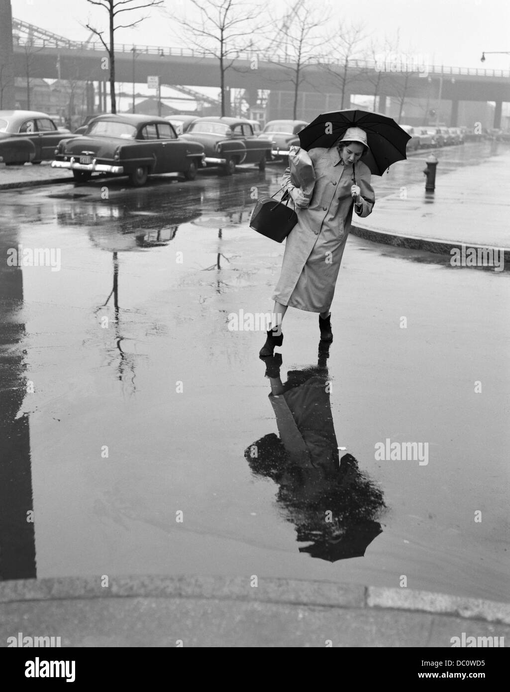 Anni Cinquanta la donna in cappotto di pioggia HAT STIVALI HOLDING OMBRELLO STEPPING BORSETTA PER EVITARE pozzanghere durante l attraversamento strada di città in fallo METEO Foto Stock