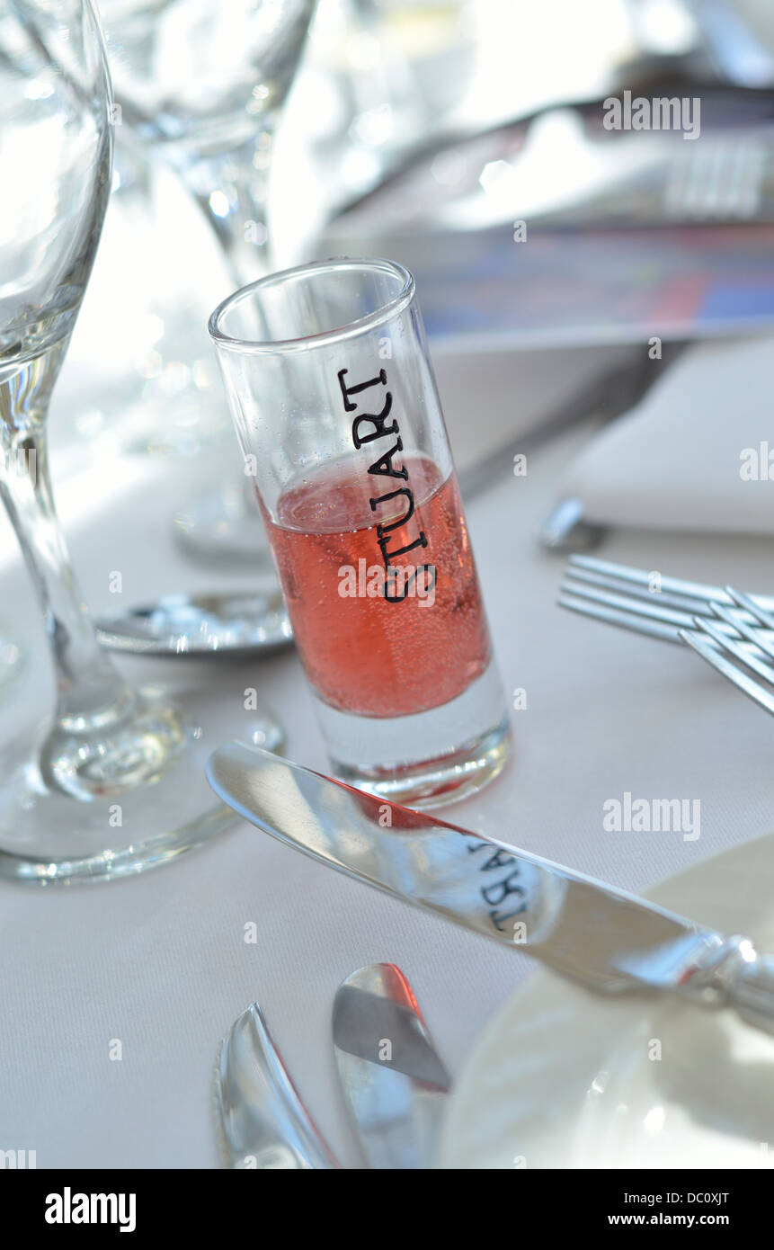 Colpo di vetro bomboniera favore rosa contenente liquido frizzante con la parola Stuart sul vetro circondato da posate su un tavolo Foto Stock