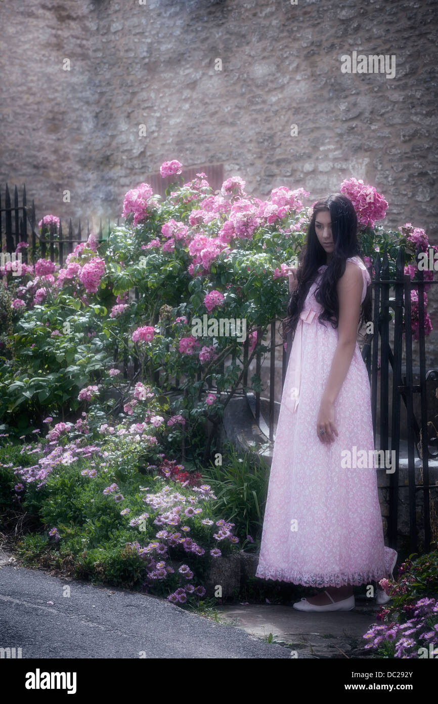Una bella donna con lunghi capelli neri in piedi in un vintage abito rosa di fronte a un cancello con fiori Foto Stock