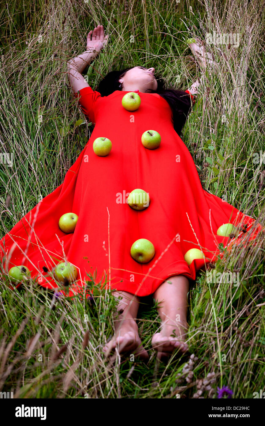 Una ragazza in un abito rosso, giacente in erba con mele verdi sul suo vestito Foto Stock