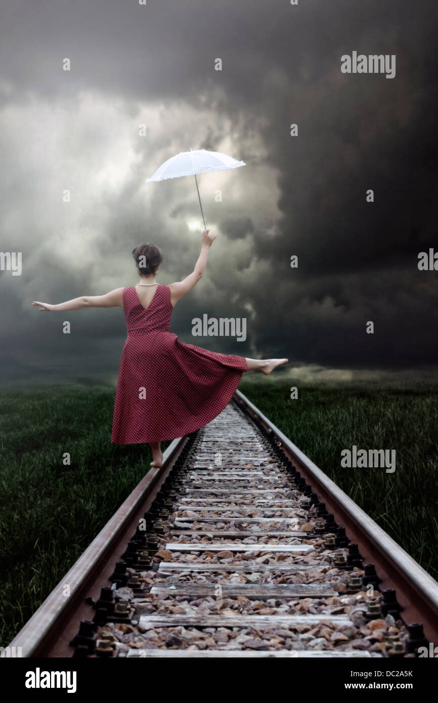 Una ragazza in un abito rosso è in equilibrio su binari ferroviari con un ombrellone bianco Foto Stock