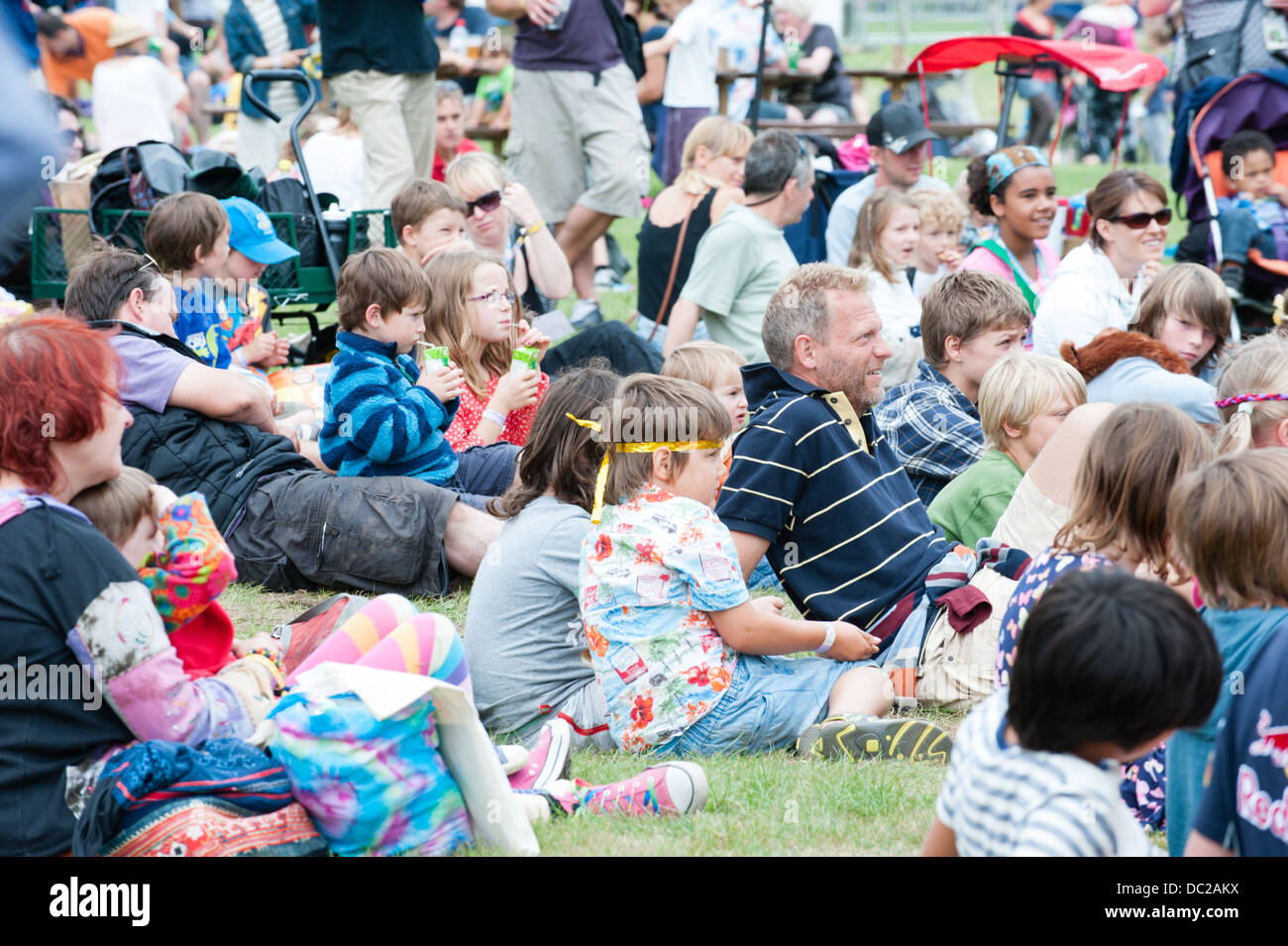 Una folla di bambini felici, adulti e famiglie seduto sull'erba godendo di un festival estivo di guardare un animatore di bambini Foto Stock