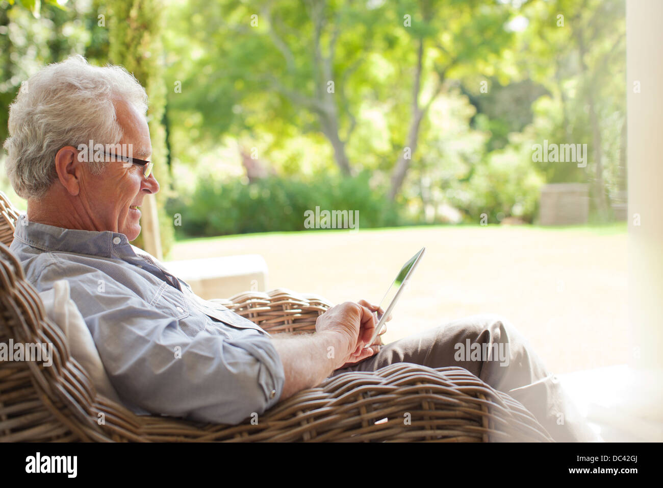 Senior uomo con tavoletta digitale in poltrona di vimini sul portico Foto Stock