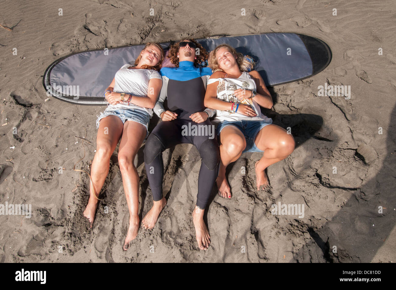 Due ragazze e guy recante a bordo sacco sulla spiaggia di sabbia nera , Ngarunui Beach, Raglan, Nuova Zelanda Foto Stock