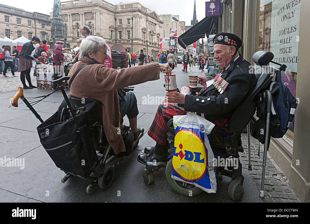 Thomas Gilzean.Mobility scooter. Disabilità. lady sulla mobilità scooter mettendo i soldi in una scatola di beneficenza tenutasi il mio uomo in uniforme in sedia a rotelle. Foto Stock