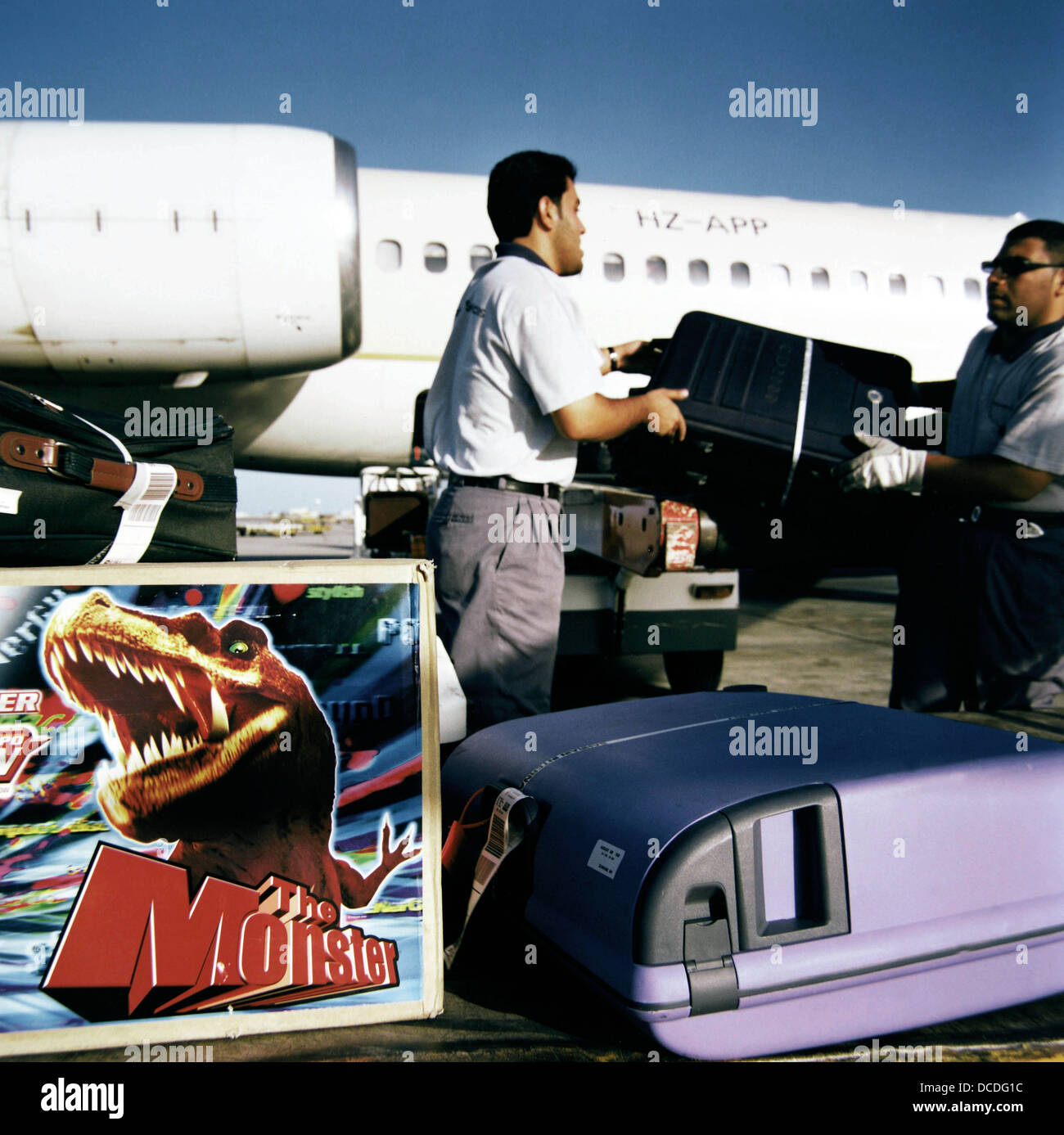 Con le valigie e un dinosauro toy box in primo piano, due bagagli manhandle gestori di sacchi su un saudita Compagnie aeree jet. Foto Stock