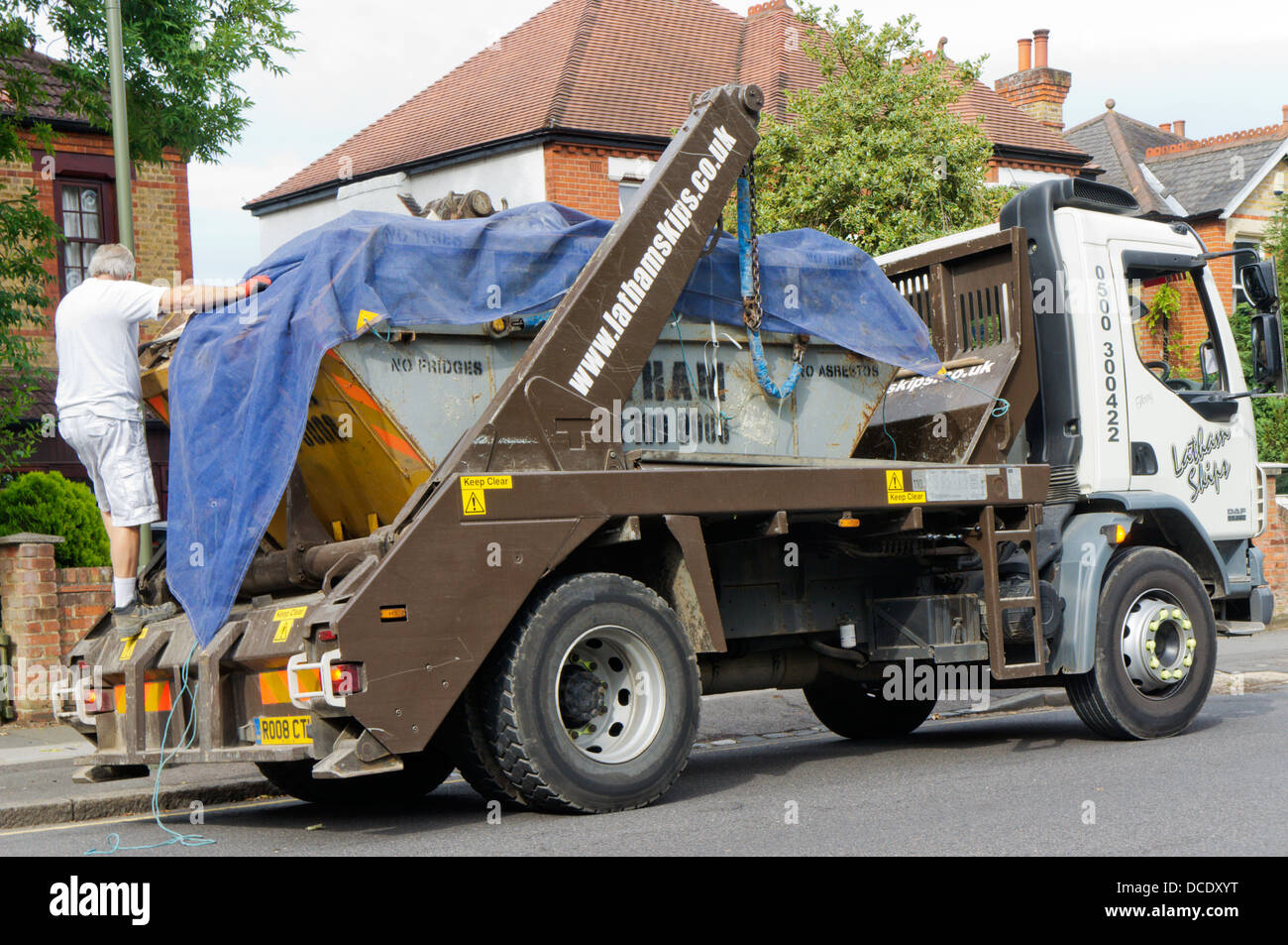 Driver carico di copertura su skip camion con telone foglio in una strada suburbana. Foto Stock