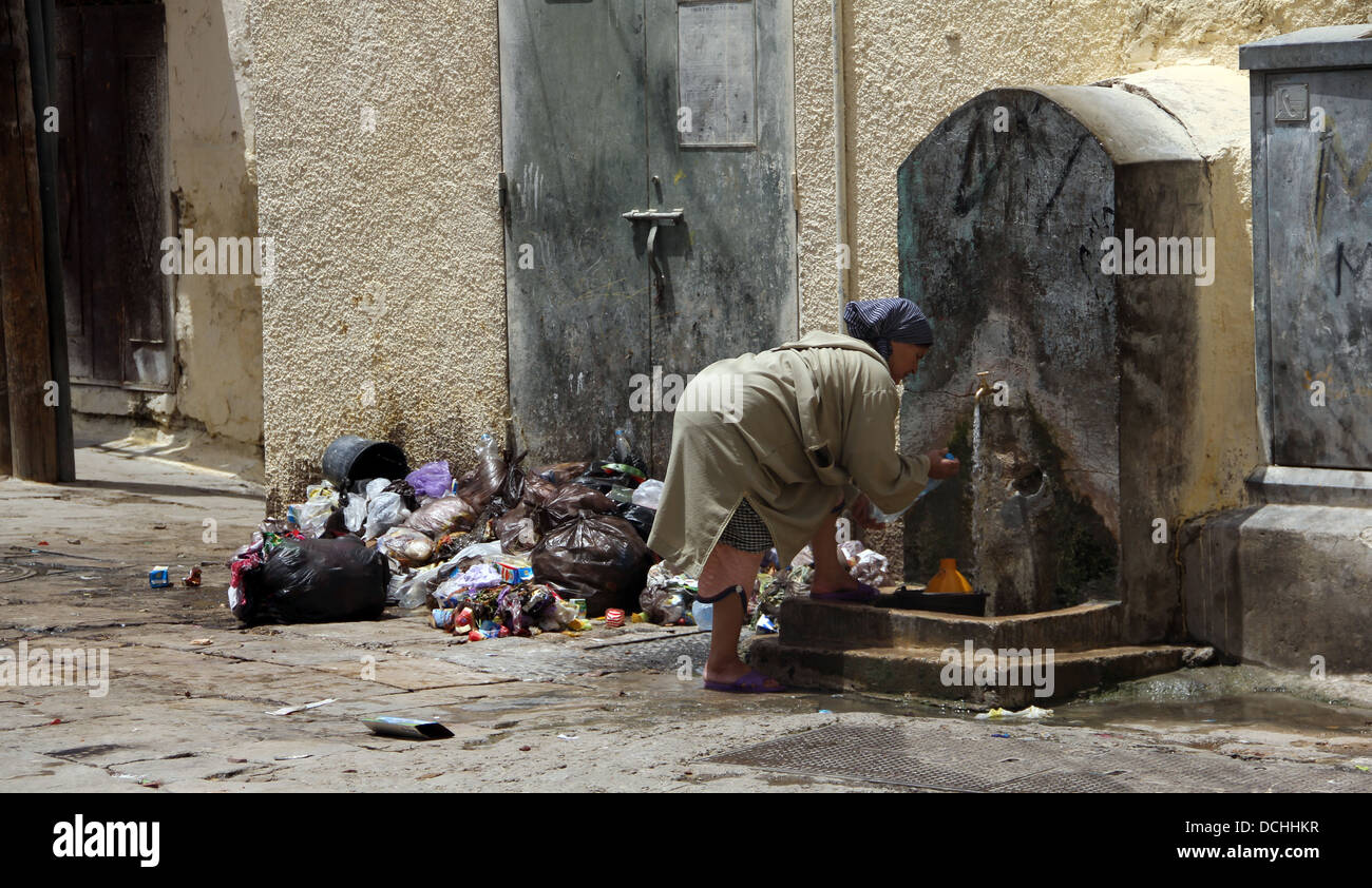 Una donna lava le mani da una fontana accanto a un mucchio di rifiuti nella medina di Fez. Foto Stock