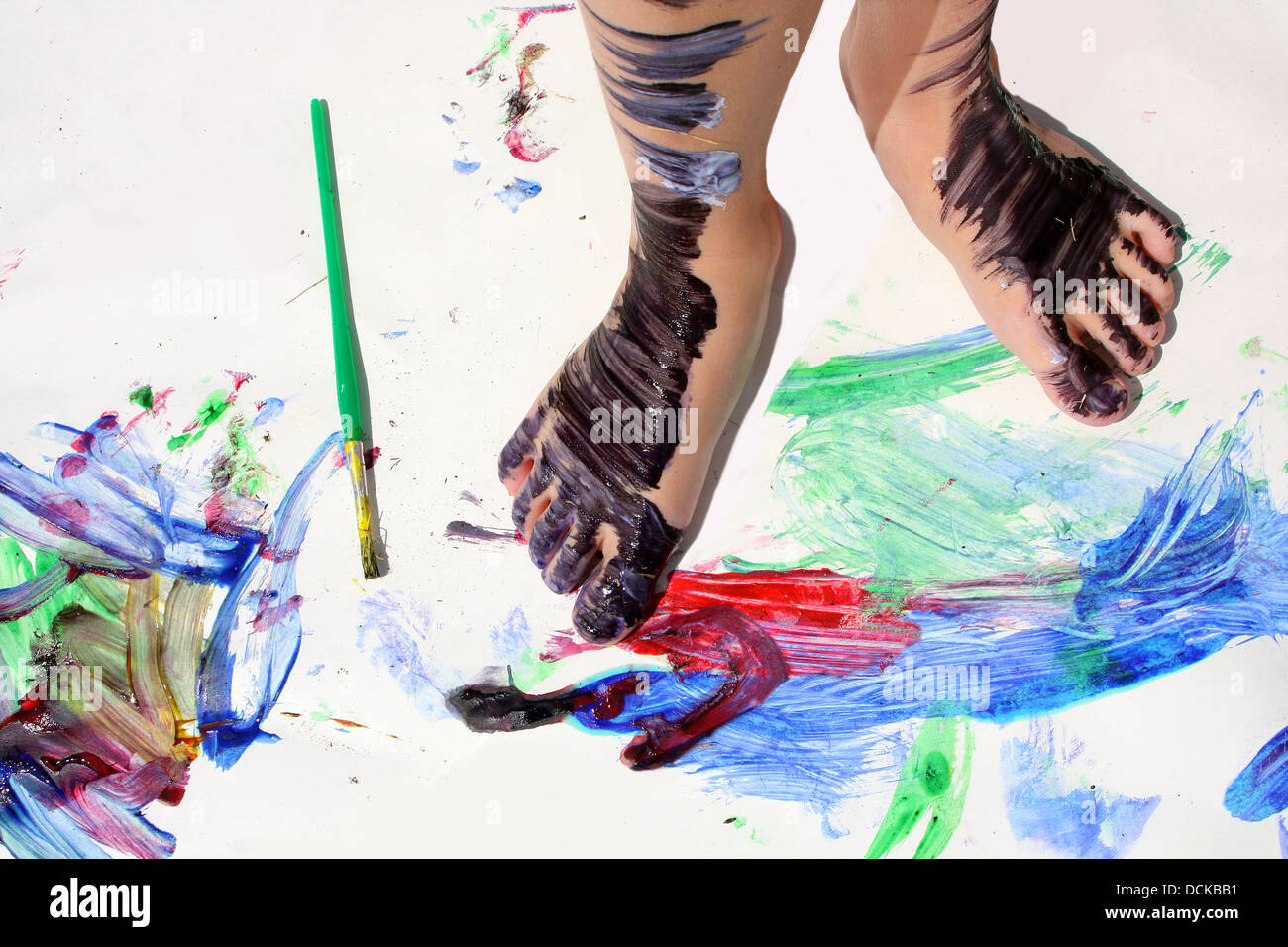 Un giovane bambino i piedi sono coperti di vernice colorata come essi sono in piedi su un pezzo di carta bianco coperto di vernici luminose Foto Stock