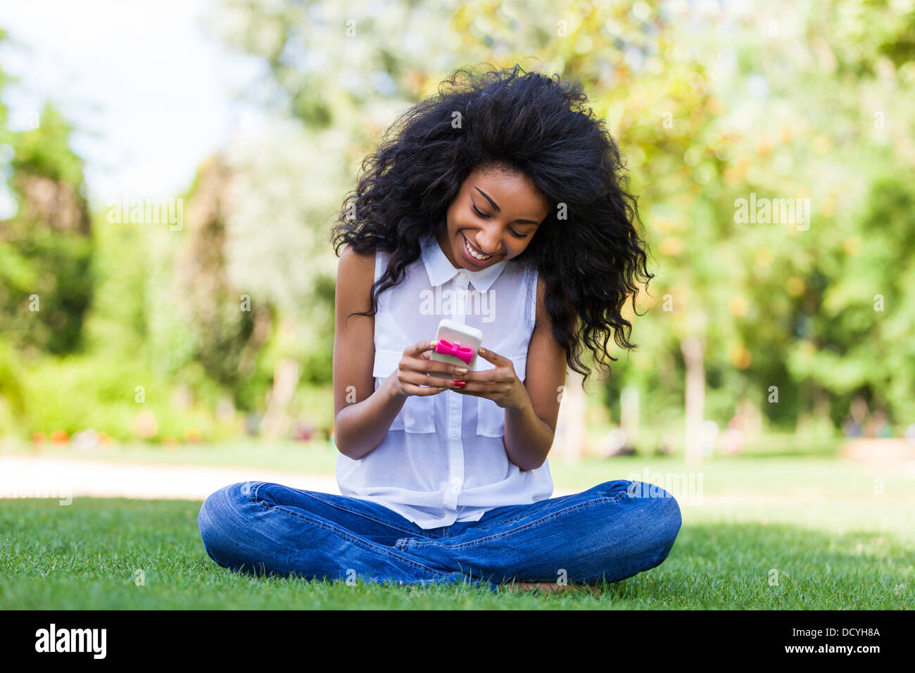 Adolescente sorridente ragazza nera utilizzando un telefono, seduto sull'erba - popolo africano Foto Stock