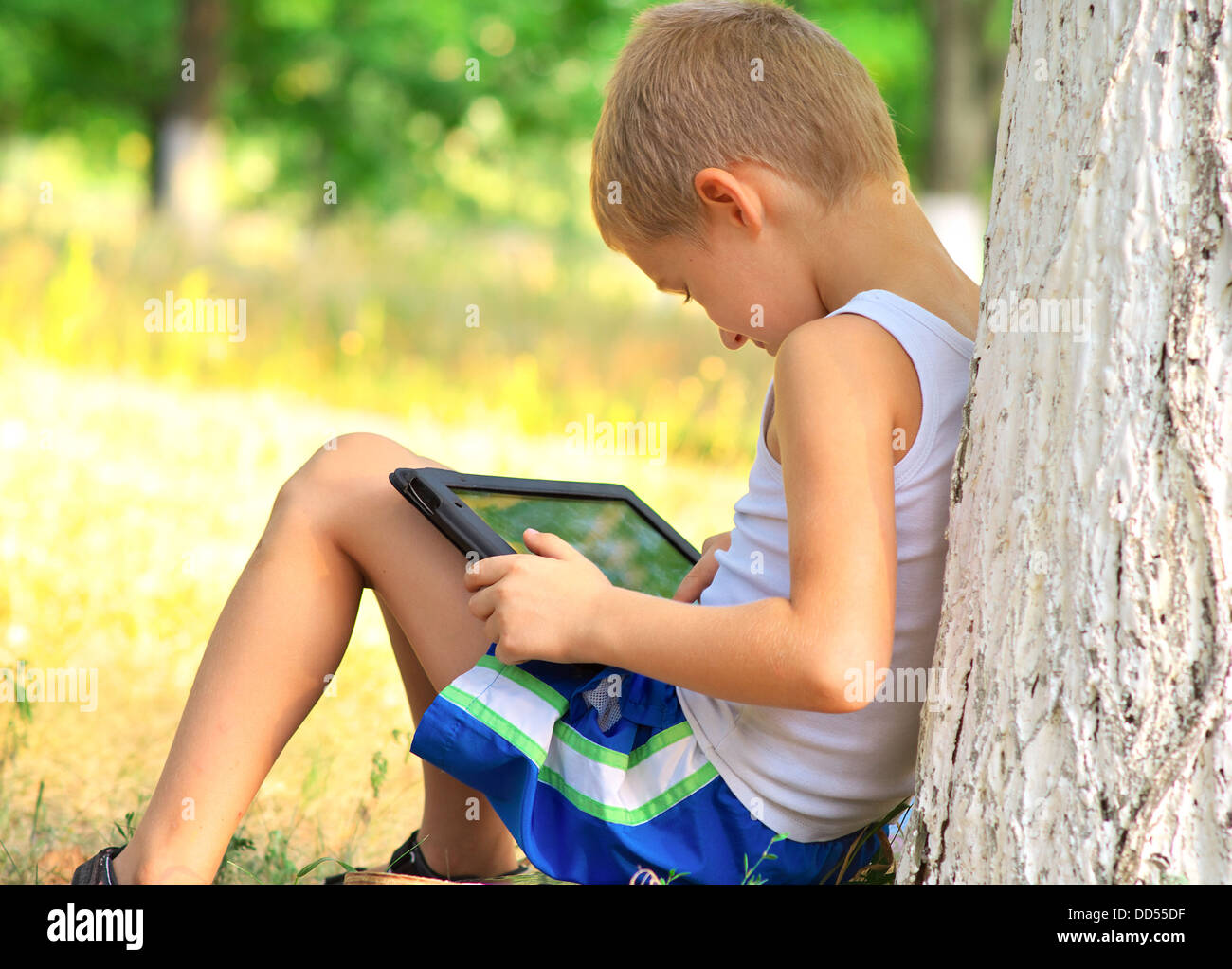 Ragazzo bambino che gioca con i Tablet PC seduto su skateboard Outdoor con bosco su sfondo Dipendenza dal Gioco di concetto Foto Stock