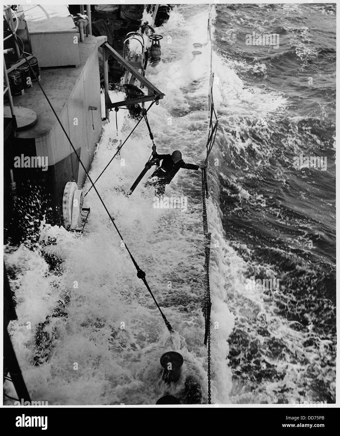 Un guardiano della costa degli Stati Uniti si vede afferrare un tubo del carburante e una linea di vita della catena, lottando per mantenere la sua posizione durante una impegnativa operazione di salvataggio in mare. L'immagine evidenzia la natura pericolosa dei salvataggi marittimi e le esigenze fisiche del personale della Guardia Costiera. Foto Stock