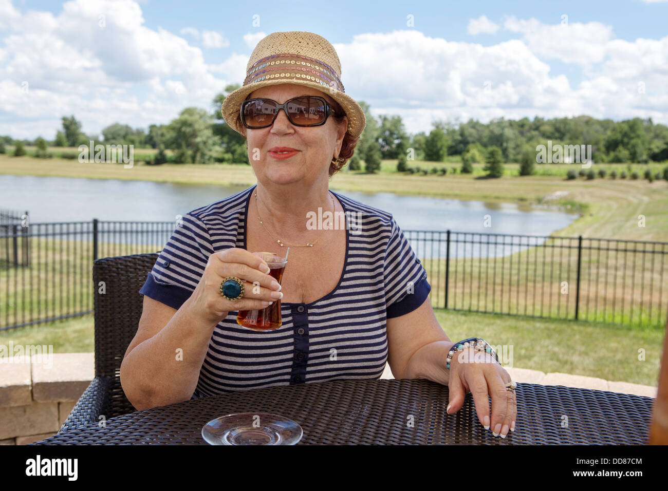 Elegante donna senior in un cappello e occhiali da sole godendosi un drink all'aperto seduti ad un tavolo con vista sul lago Foto Stock
