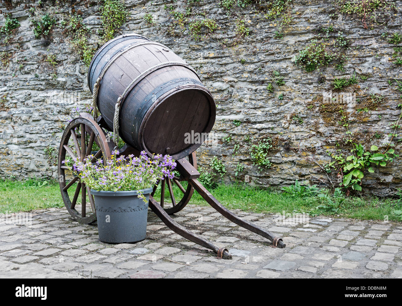 Vecchia botte di vino con ruote e parete con rocce e fiori Foto Stock